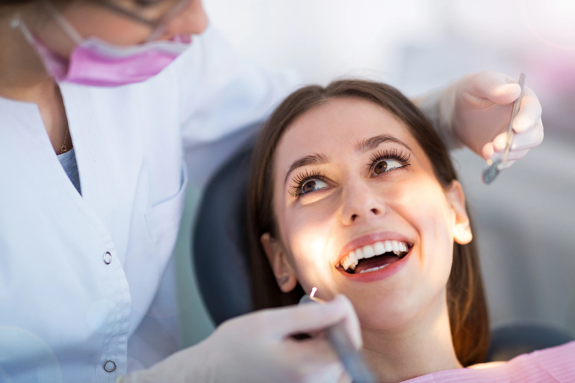 Dentist in a white coat and pink mask performs a dental exam on a smiling patient under bright, clinical office lighting.