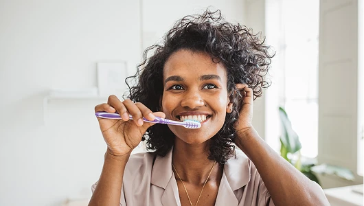 Smiling woman with curly hair brushing teeth with a purple toothbrush in a bright, sunlit room with a modern minimalist aesthetic.