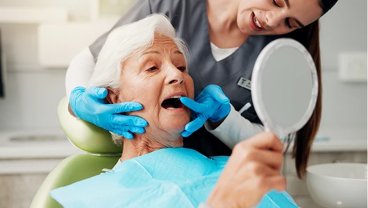 Dental professional in grey scrubs examining a senior patient’s teeth. Patient holds a mirror in a brightly lit, sterile clinical dental office.