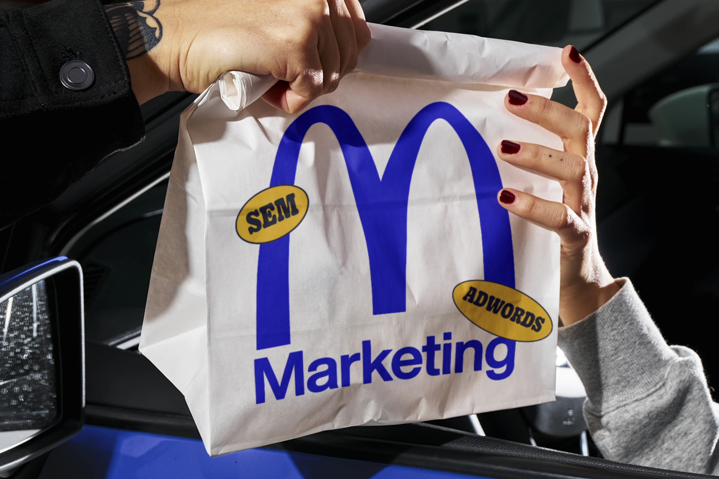 A takeaway bag getting passed through a car window, with a 'Marketing' logo on the front and SEM stickers
