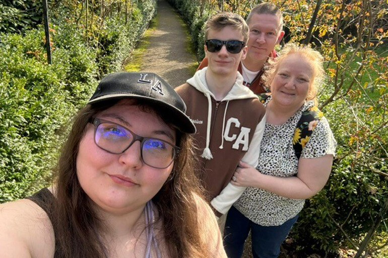 A young woman takes a selfie of herself and her family ( young man and two parents) along a leafy lane.