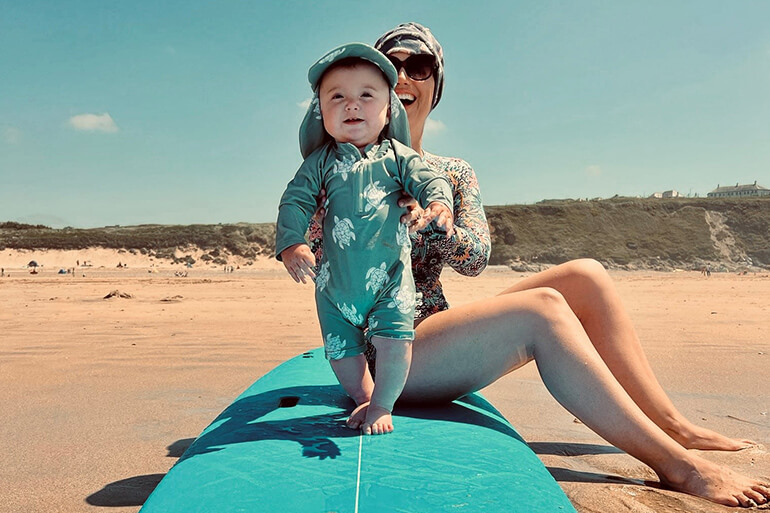 A small toddler in a sunsuit and hat is held by her mum on a beach on a sunny day. The child is standing on a soft-top surfboard whilst the mum sits on the same board behind her.