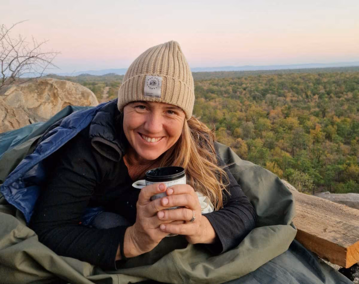 Smiling woman wearing a beige knit hat holding a cup, wrapped in a sleeping bag outdoors at sunset.