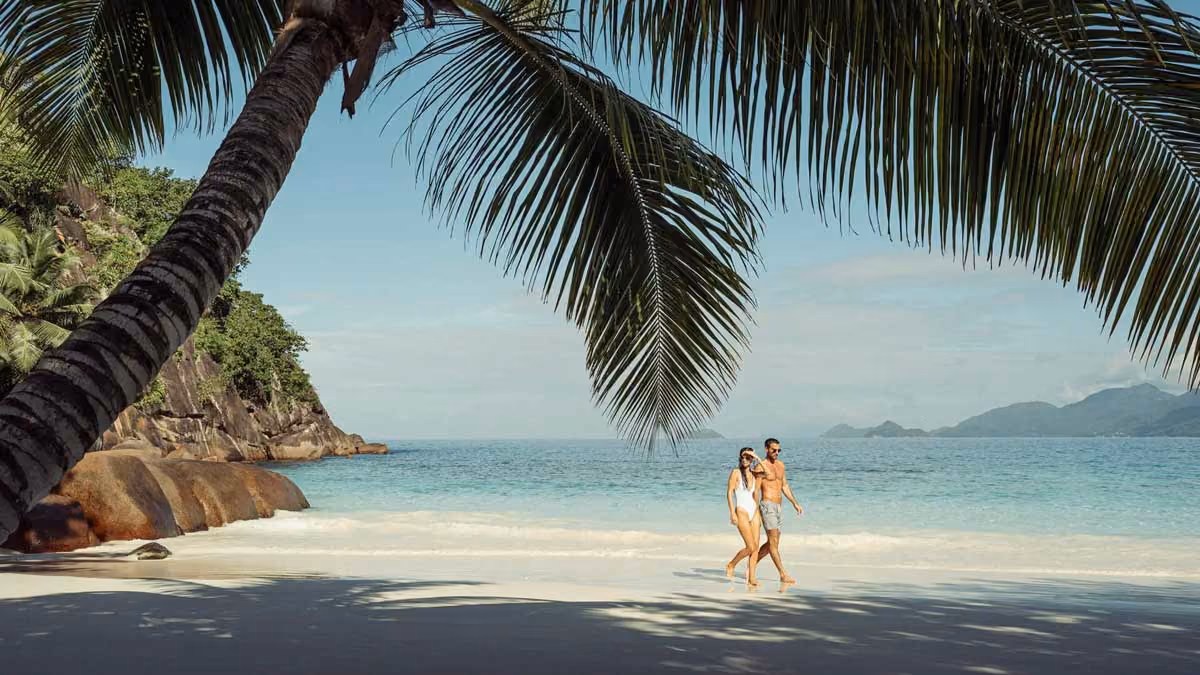 Couple walking on a sunny tropical beach under palm trees with ocean and mountains in the background.