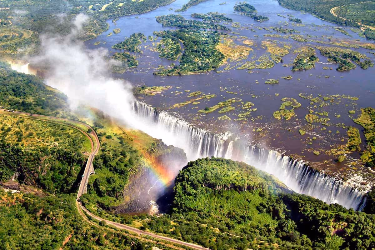 Aerial view of a large waterfall with mist, rainbows, surrounding greenery, and a winding road nearby.