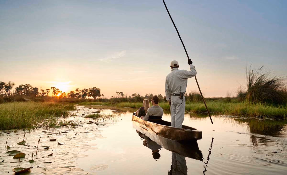Two people sitting in a canoe with a guide standing and pushing with a pole on a calm river at sunset.