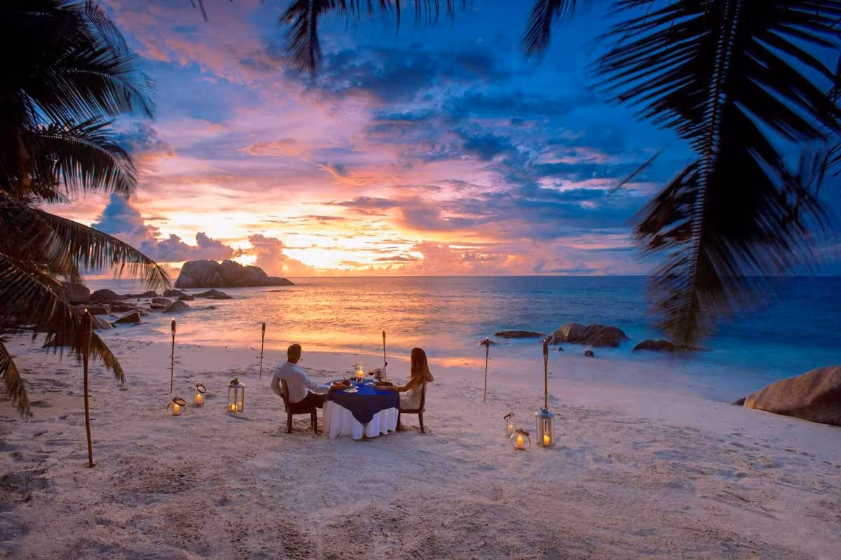 Couple dining at a table on a sandy beach at sunset, surrounded by lanterns and tiki torches.
