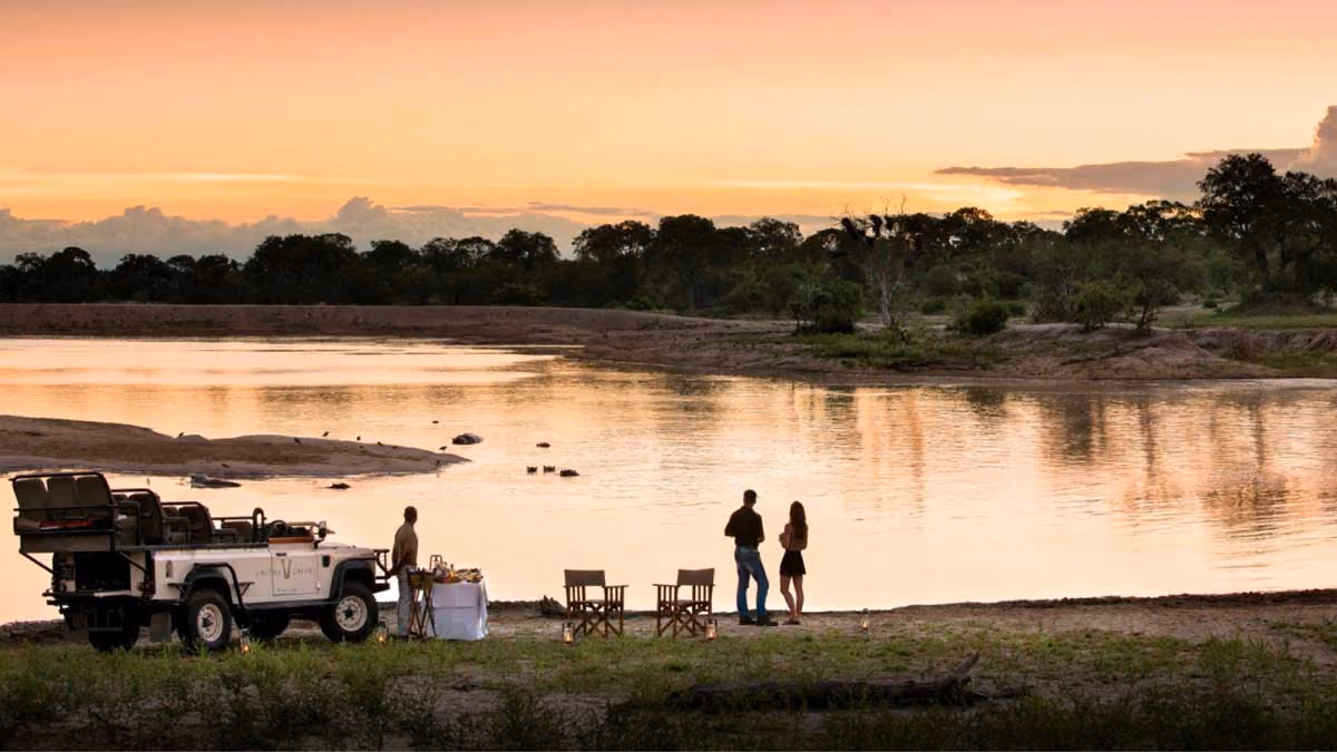 Couple by a lake at sunset with safari vehicle, table with food, and chairs in a wild setting.