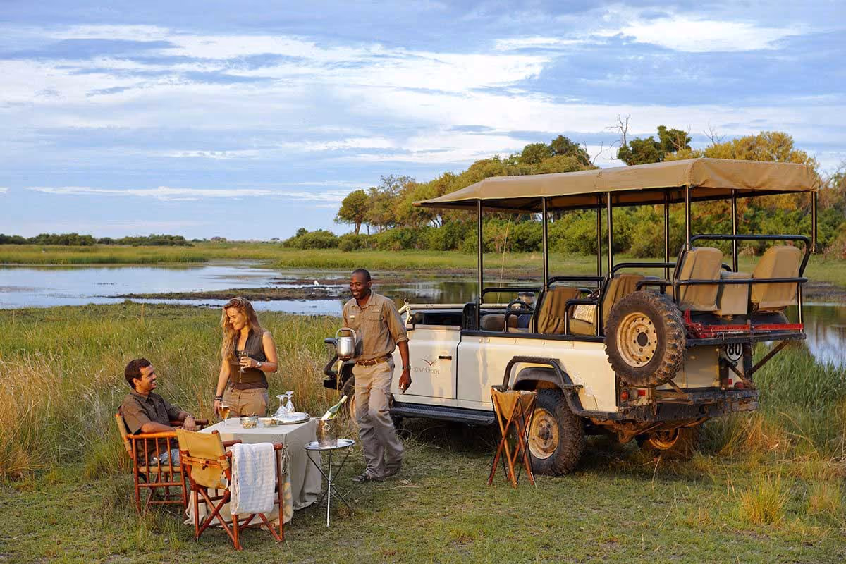 Two people seated outdoors next to a safari vehicle with a table set for drinks by a riverside.