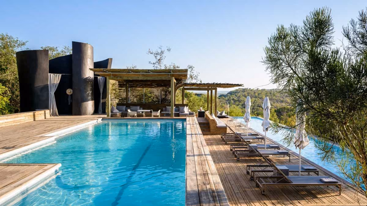 Luxury outdoor pool with sun loungers, umbrellas, shaded seating area, and scenic greenery in the background.