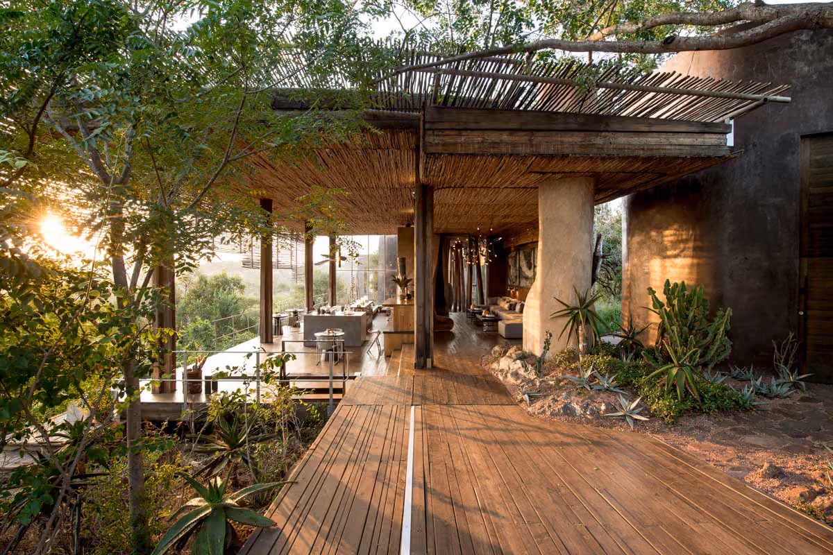 Open-air bar and lounge with wooden deck, natural plants, and a thatched roof under sunlight.