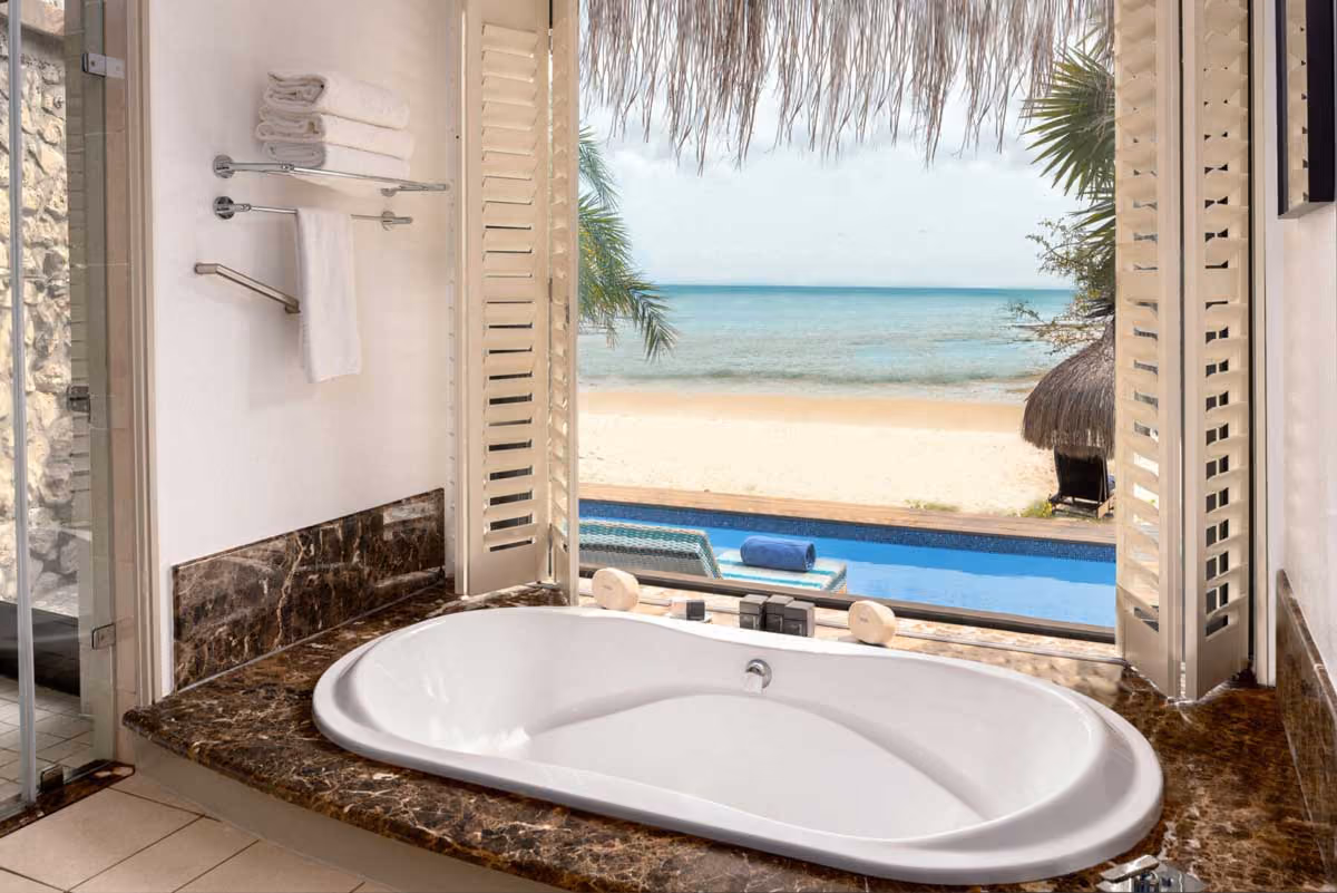 Bathroom with a large white soaking tub by an open window overlooking a pool, beach, and ocean view.