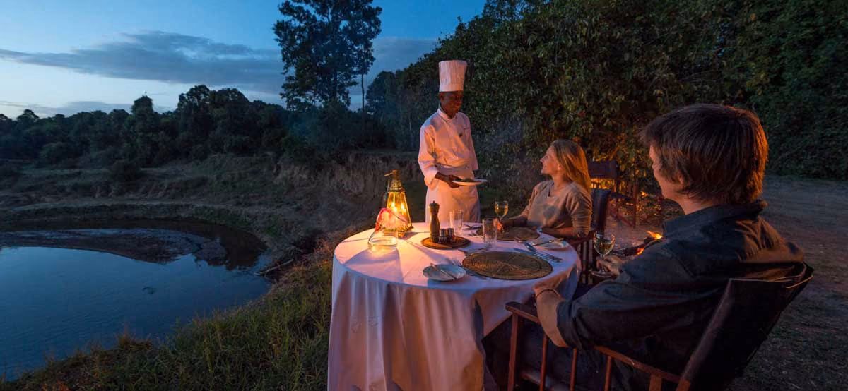 Couple enjoying outdoor dinner at dusk served by chef near a pond surrounded by trees.