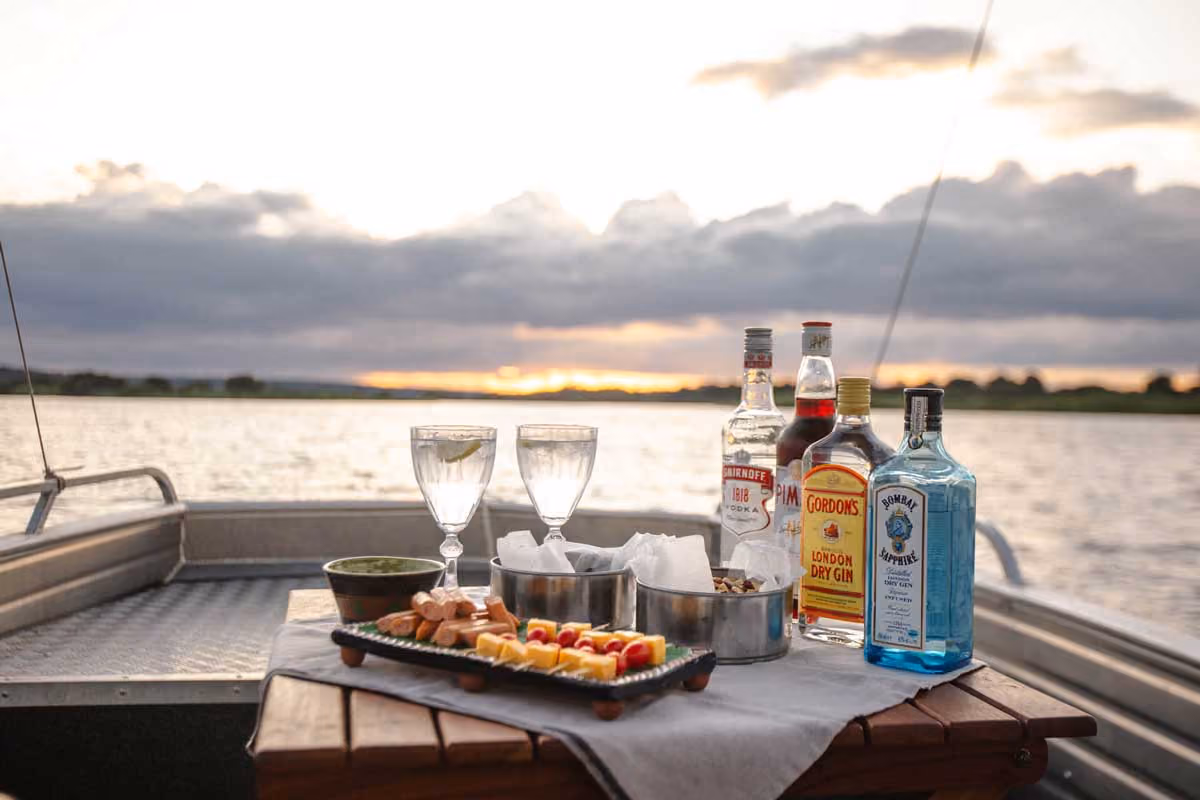 Table on a boat with bottles of alcohol, two glasses, snacks, and a sunset over the water.