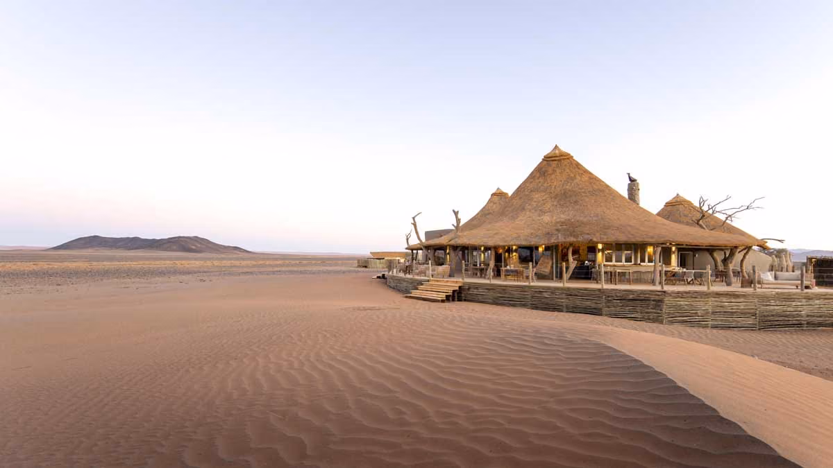 Thatched roofs building on a deck in a vast desert landscape with sand dunes and distant hills.