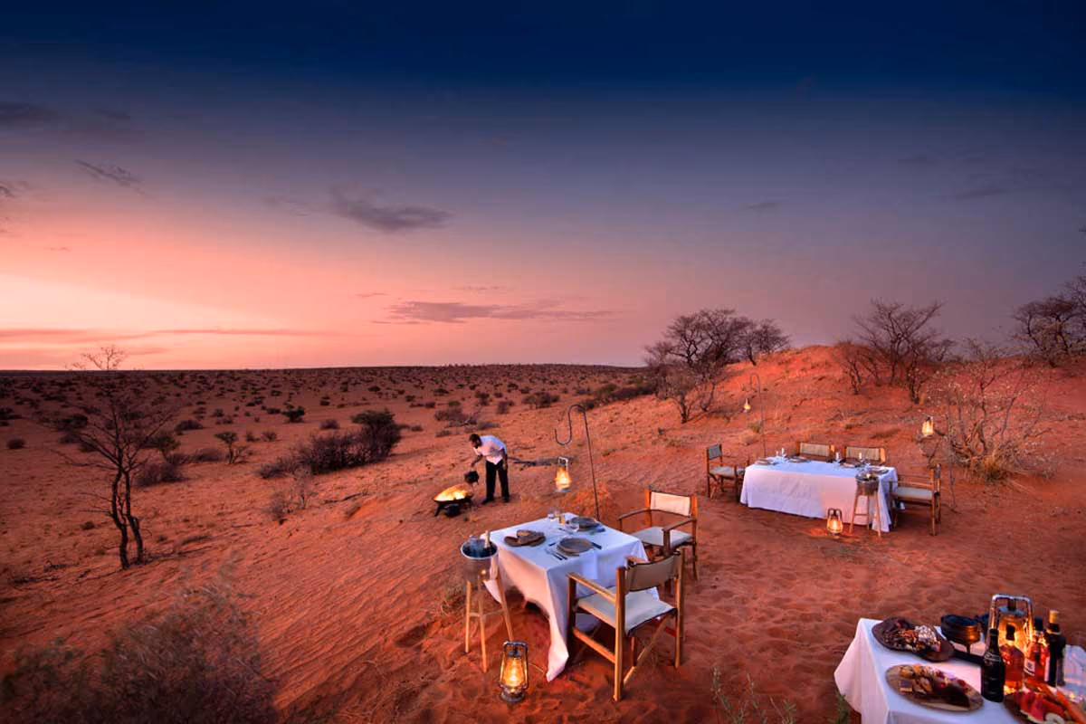 Outdoor dining setup with white tablecloths and chairs on red sand during sunset in a desert landscape.