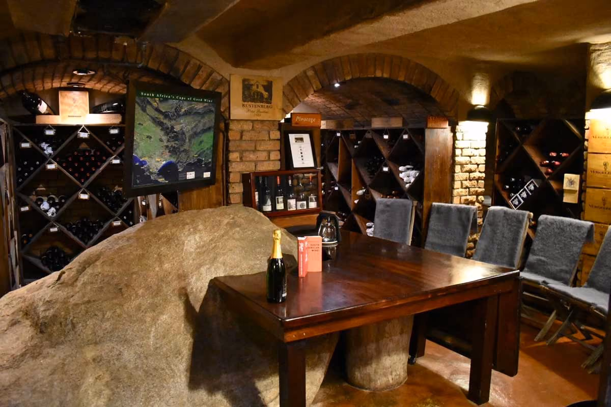 Wine cellar room with wooden racks, stone wall, table with champagne bottle, and chairs with gray covers.