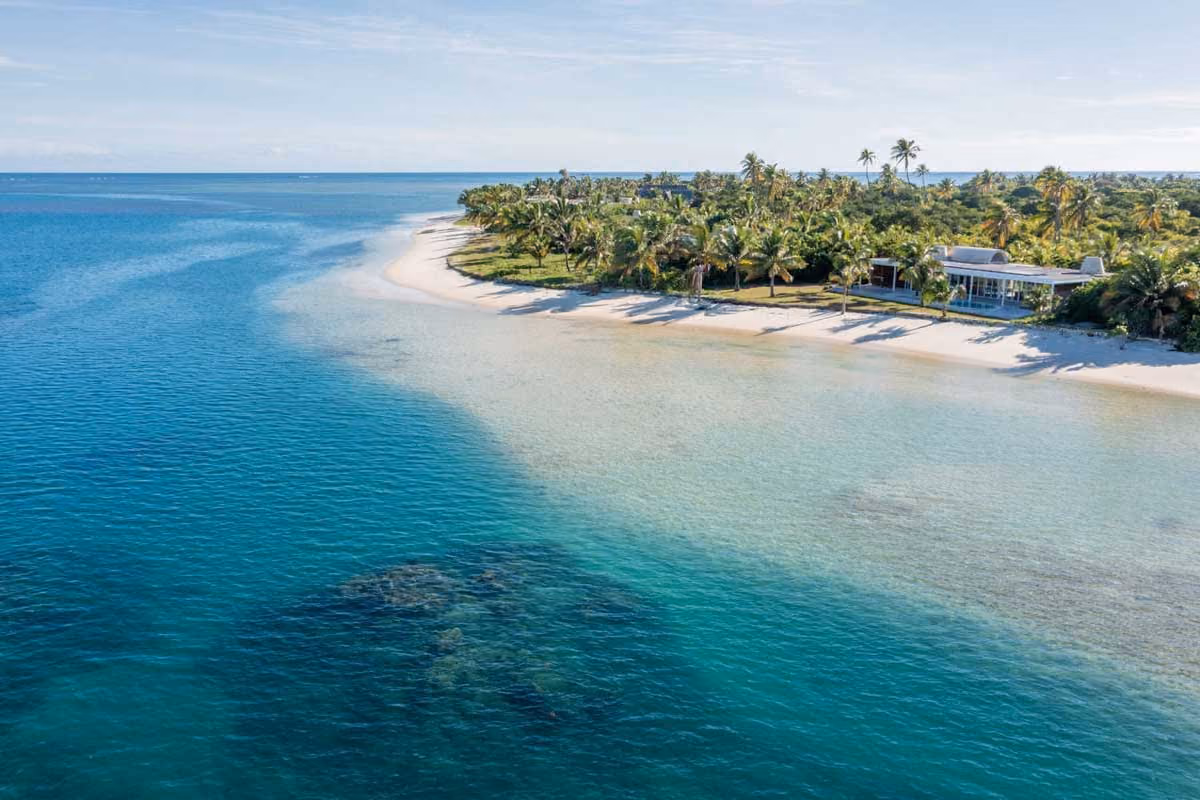 A tropical island beach with clear blue water, white sand, palm trees, and a modern house.