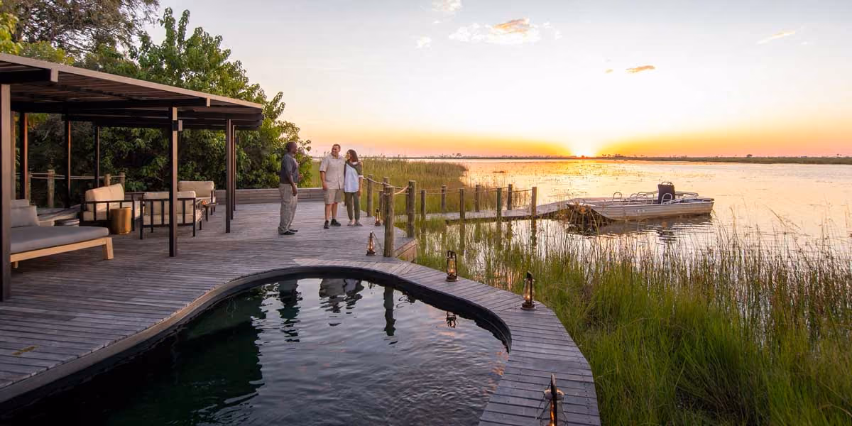 Sunset over river with docked boat beside wooden deck and three people talking near a pool.
