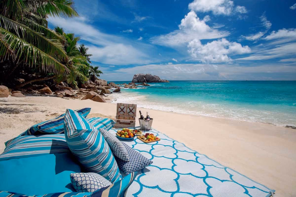 Beach setup with striped pillows, fruit plates, and drinks on a patterned mat near turquoise ocean and rocks.