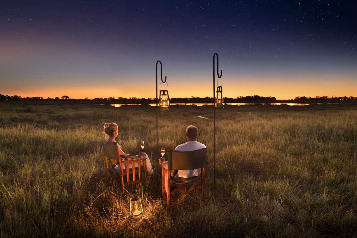 Couple sitting in wooden chairs with lanterns and wine glasses in a grassy field at sunset.