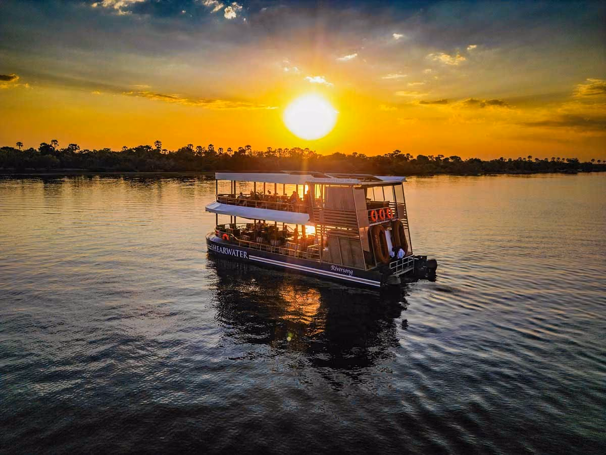 Two-level boat on calm water at sunset with sun low near horizon and silhouetted trees in background.