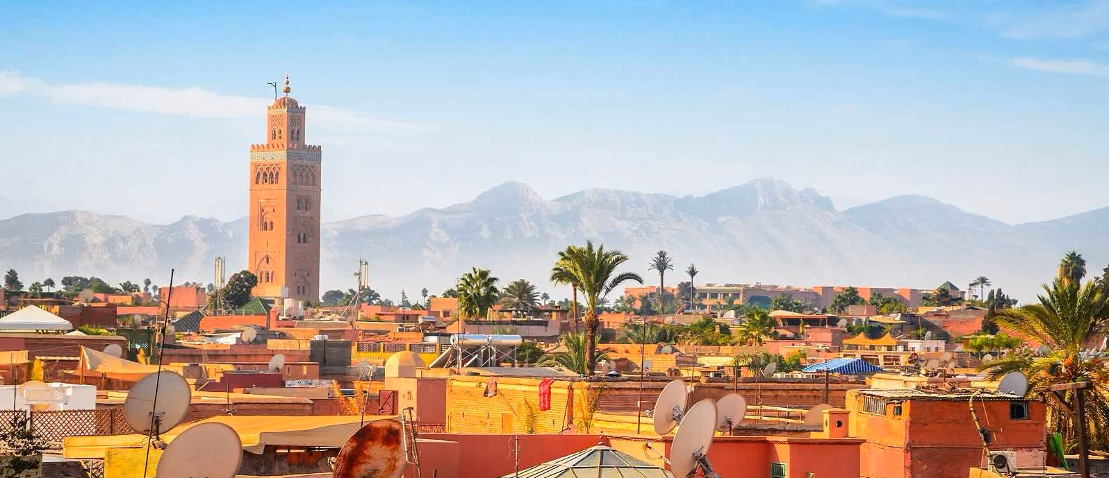 Cityscape with terracotta buildings, palm trees, satellite dishes, and a tall tower against mountain backdrop.