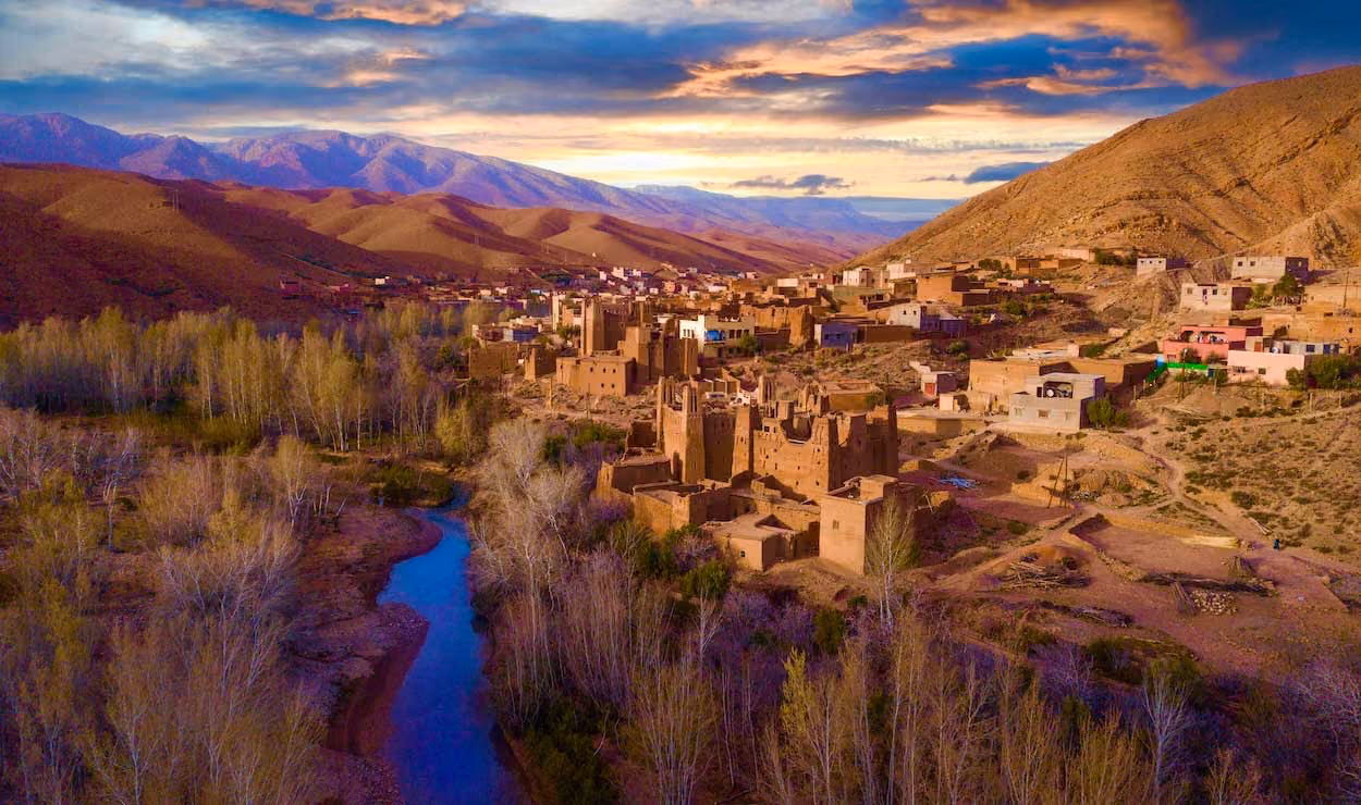 River curving through trees beside a village with brown adobe buildings in a mountainous landscape at sunset.