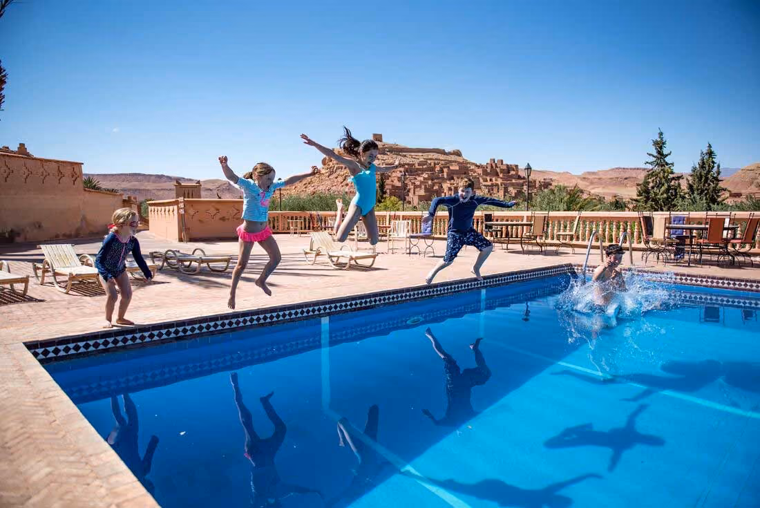 Four children jumping into an outdoor pool with splash and shadows, desert landscape in the background.