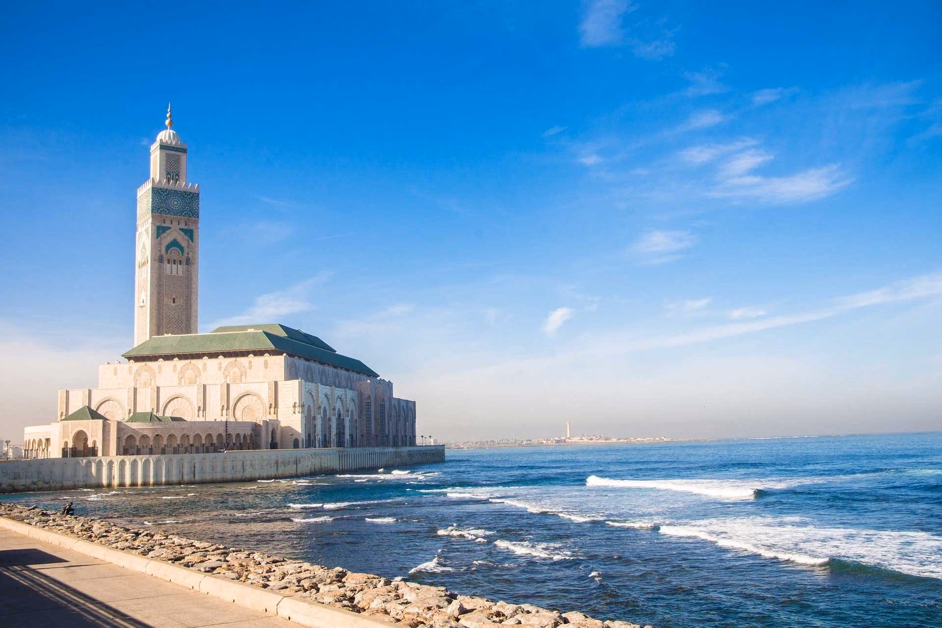 Large mosque with tall minaret by the ocean under a clear blue sky with gentle waves.