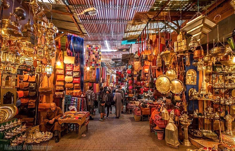 Busy market street with stalls selling lanterns, bags, brassware, and textiles under a corrugated roof.