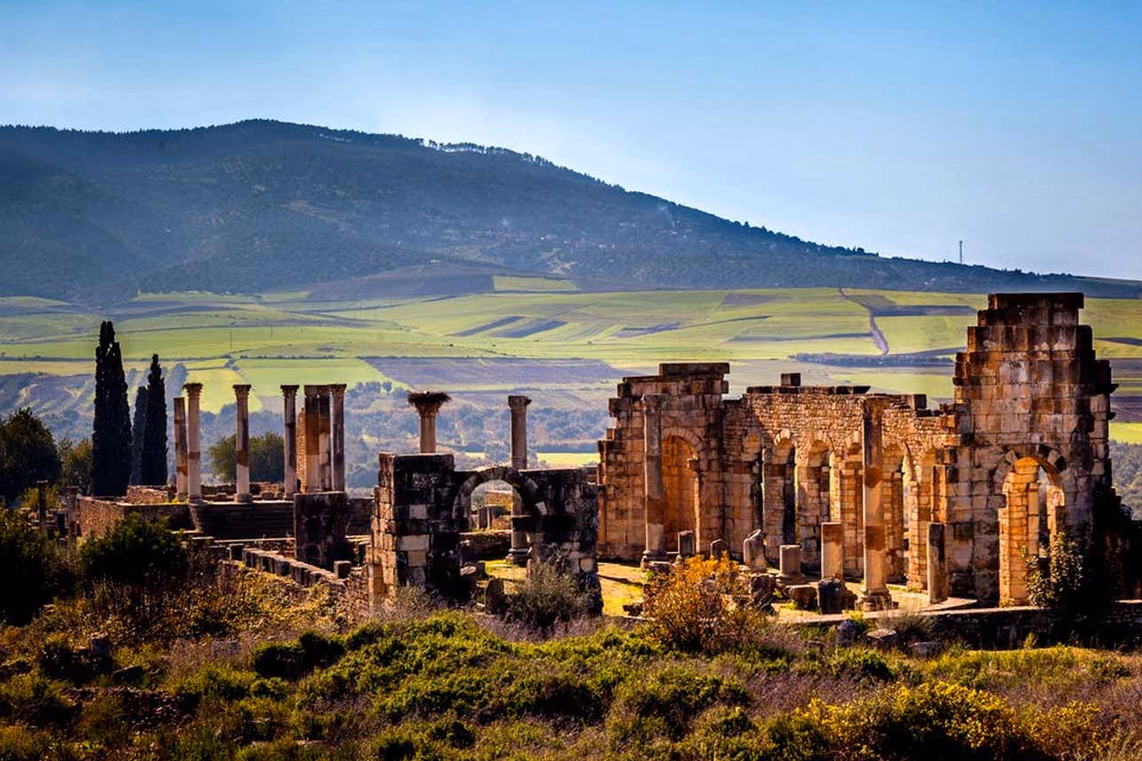 Ancient stone ruins with columns in a green valley under a clear blue sky and distant hills.