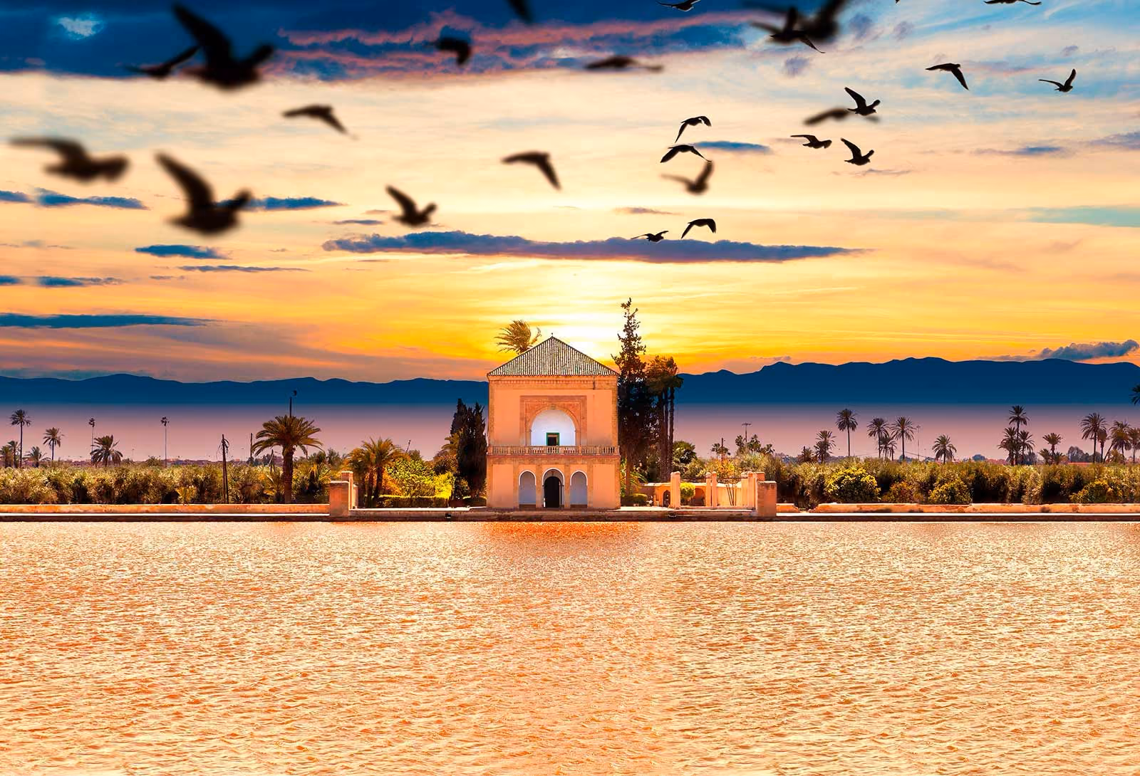 Sunset over a pavilion by a reflective pool with palm trees and mountains in the background and birds flying above.