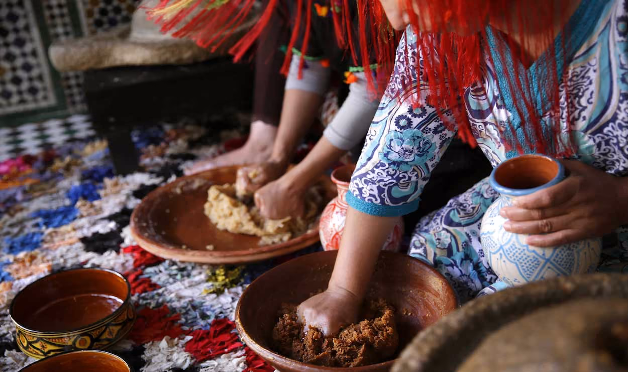 Two people mixing ingredients by hand in large bowls seated on a colorful rug with pottery nearby.