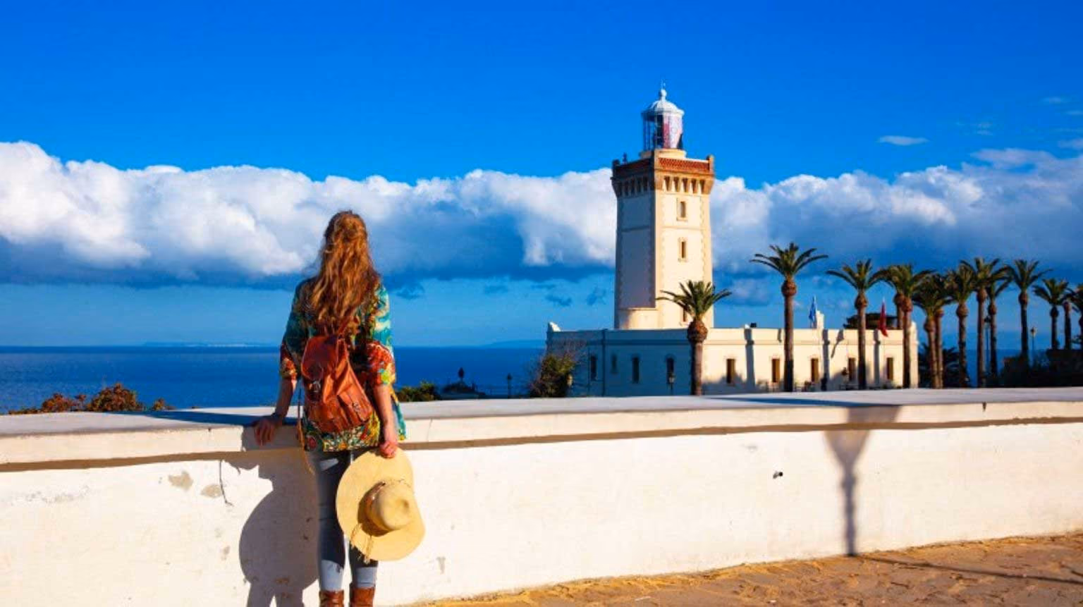 Woman with hat and backpack looking at lighthouse and palm trees under a blue sky with clouds.