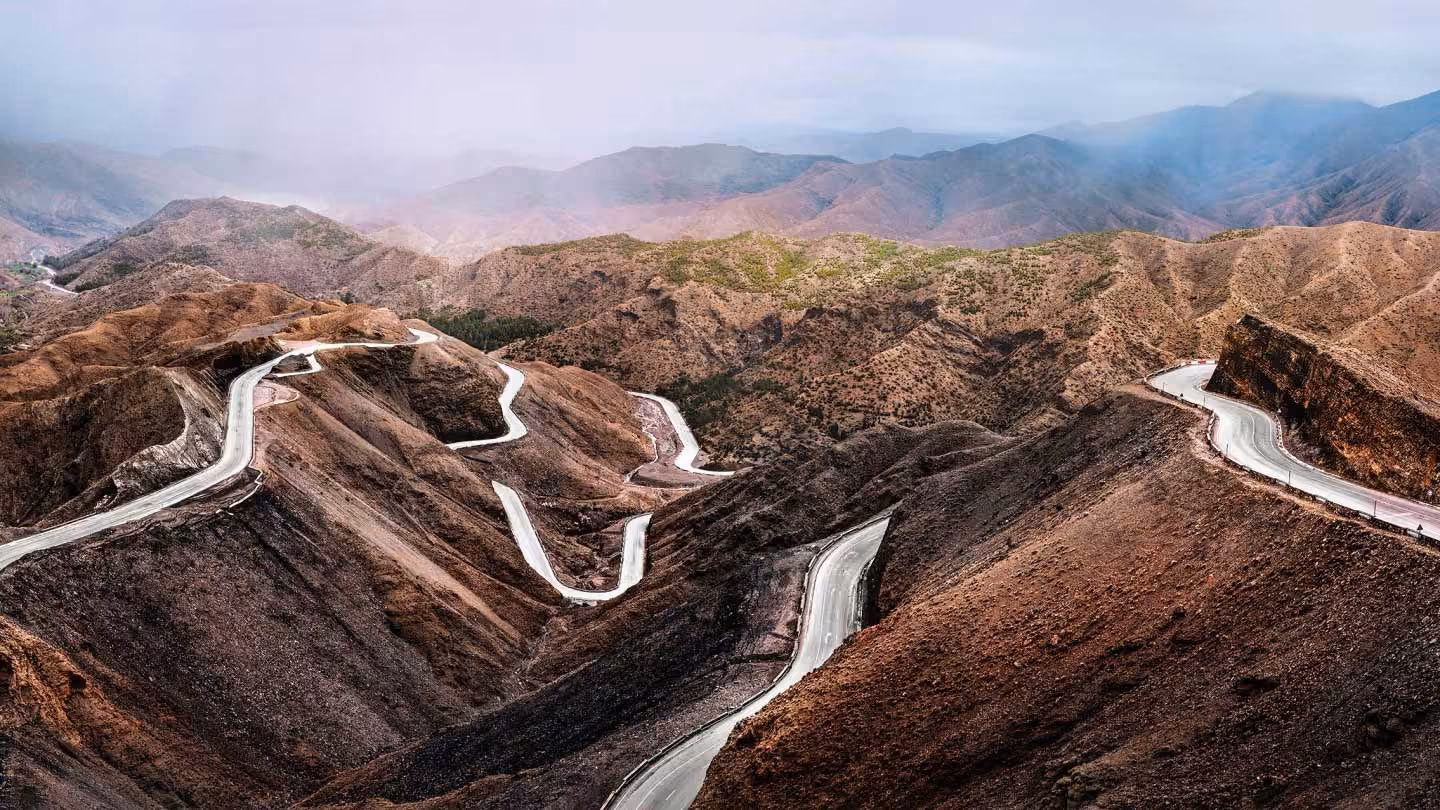Winding paved road snakes through dry, rugged mountainous terrain under a hazy sky.