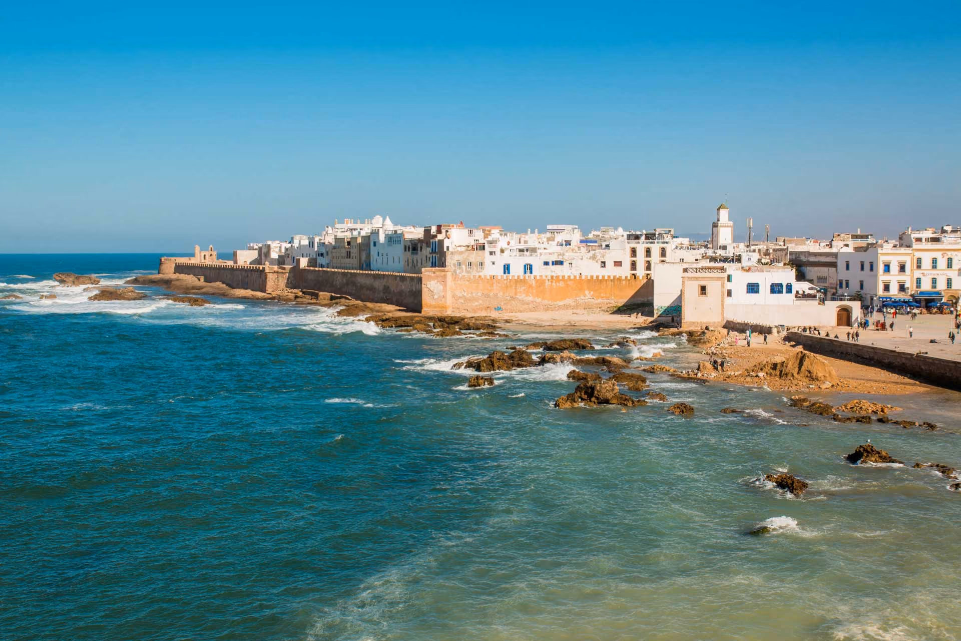 Seaside city with white buildings and stone walls along a rocky coast under a clear blue sky.