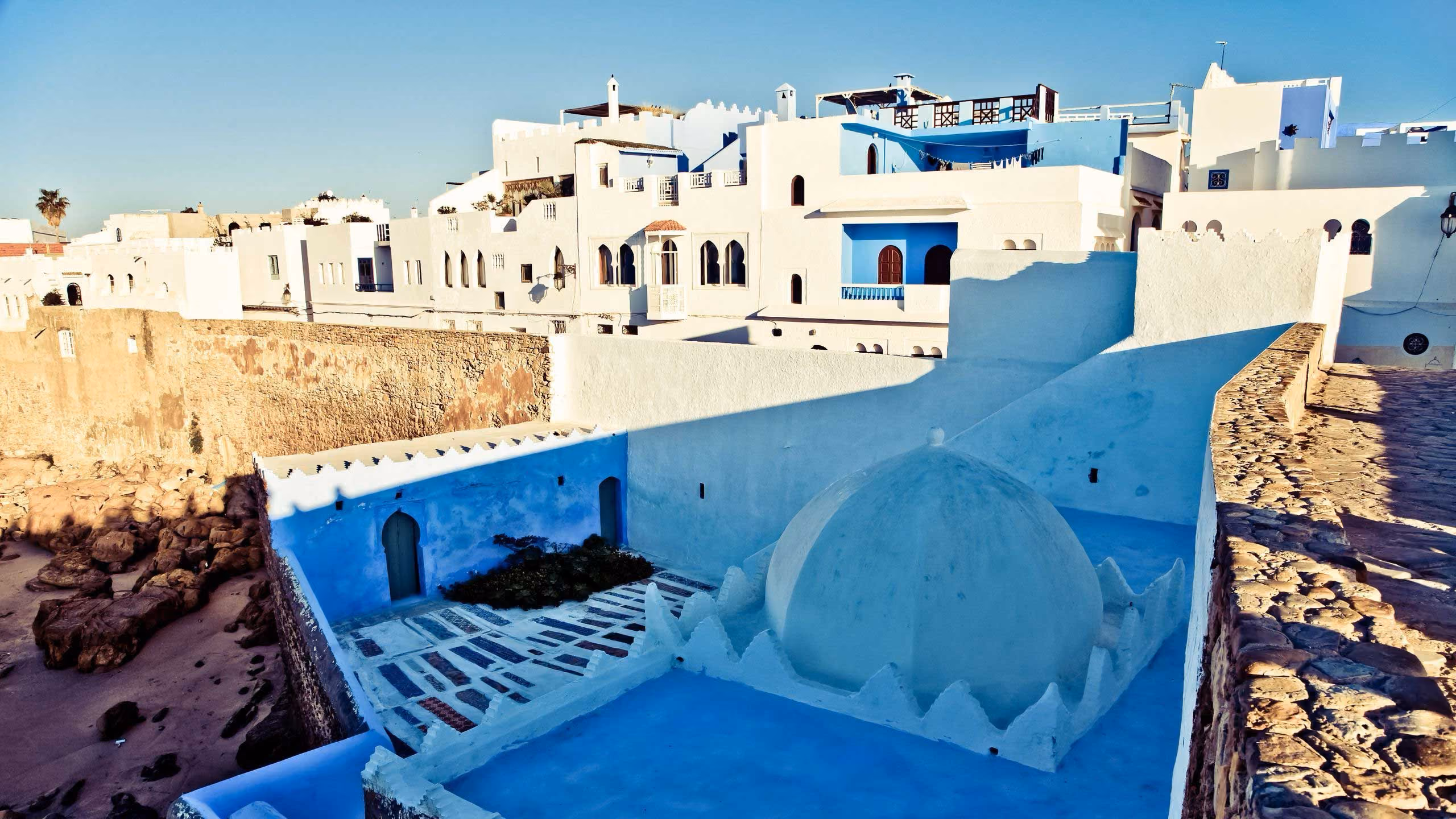 White and blue Mediterranean buildings on a rocky coastline under clear sky.