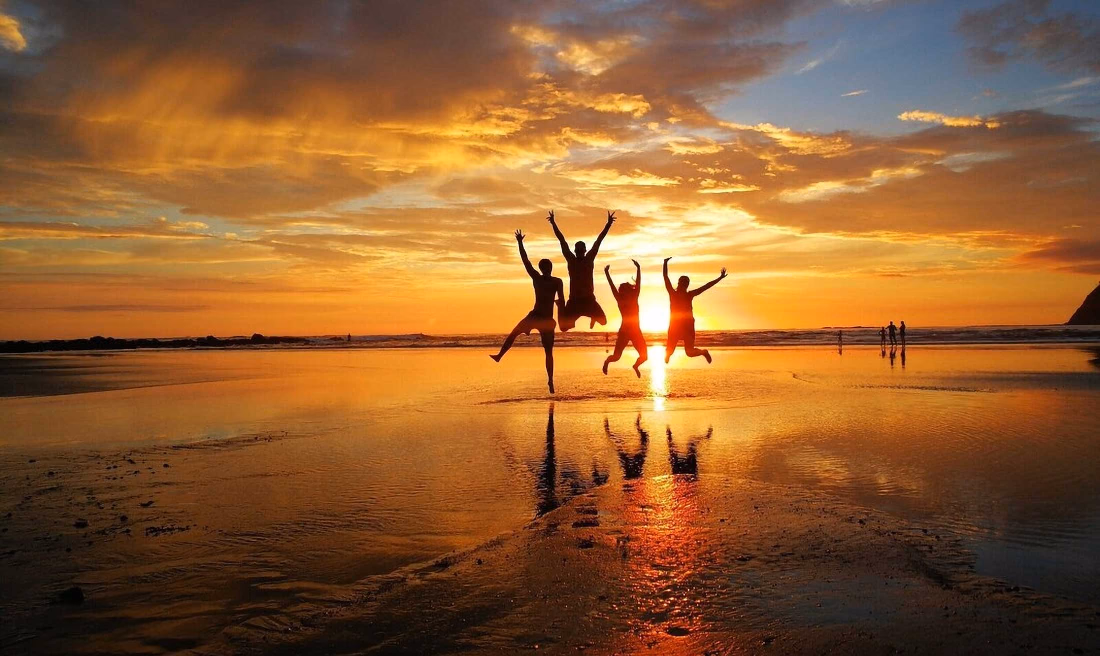 Four people joyfully jumping on a beach at sunset with their reflections in wet sand.