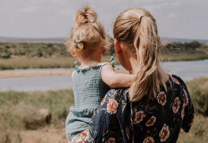 Woman with blonde hair in a ponytail holding a toddler with a small ponytail near a river and greenery.