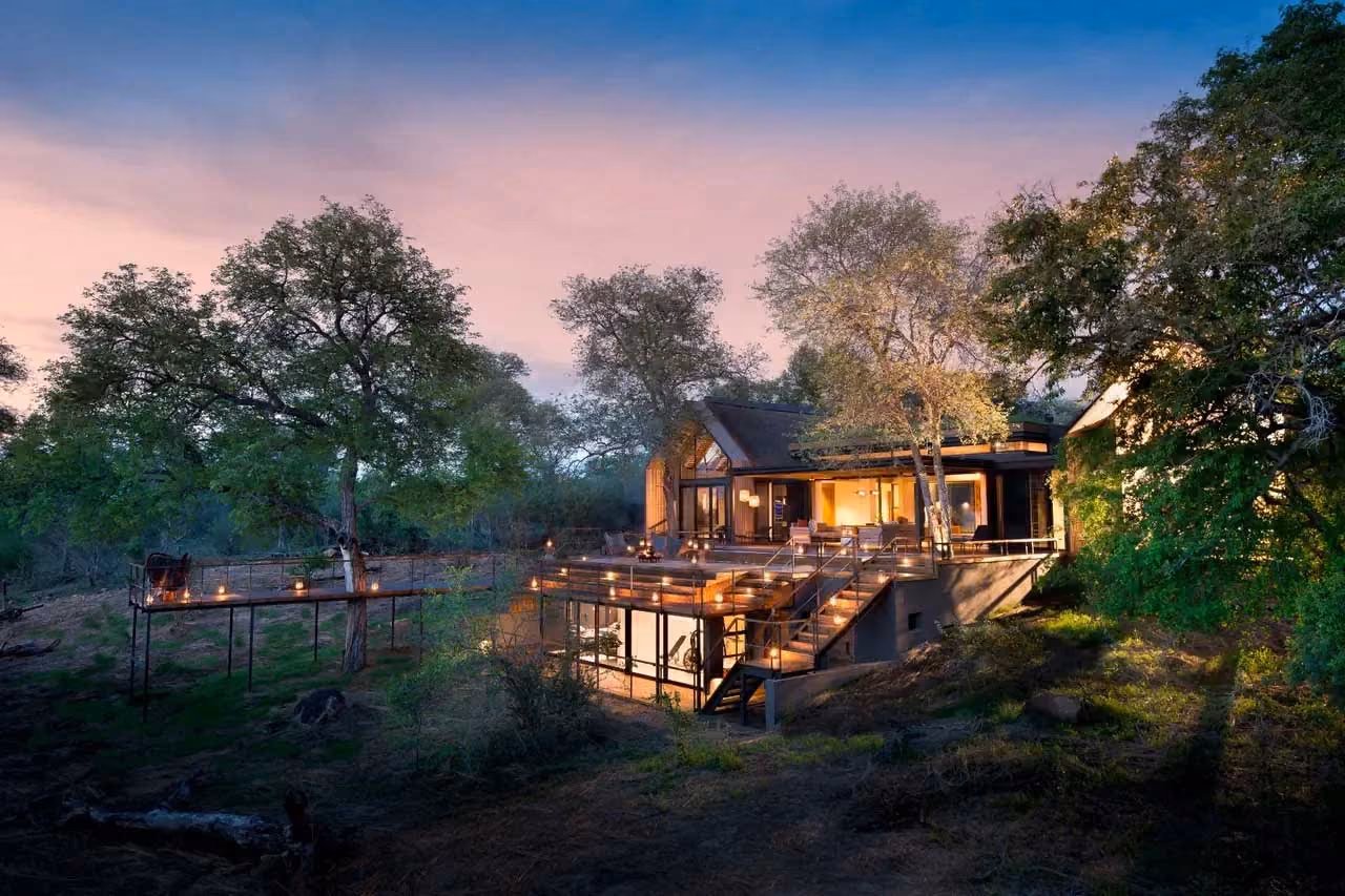 Modern house with large windows and lit deck surrounded by trees at dusk.