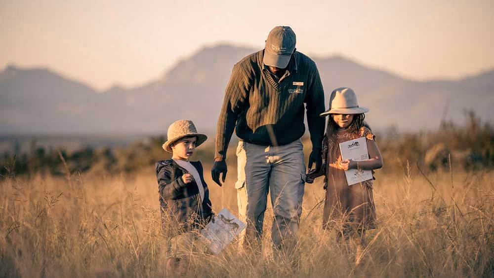 Man in cap walks with two children holding papers in a tall grass field with mountains in background.