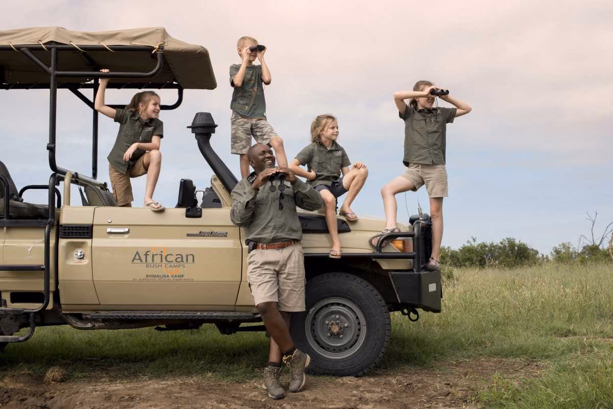 Group of kids and man in safari clothes on and around a safari truck using binoculars in grassy savannah.