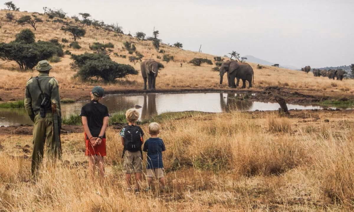 A guard and three children watch elephants near a waterhole in a dry, grassy savanna landscape.