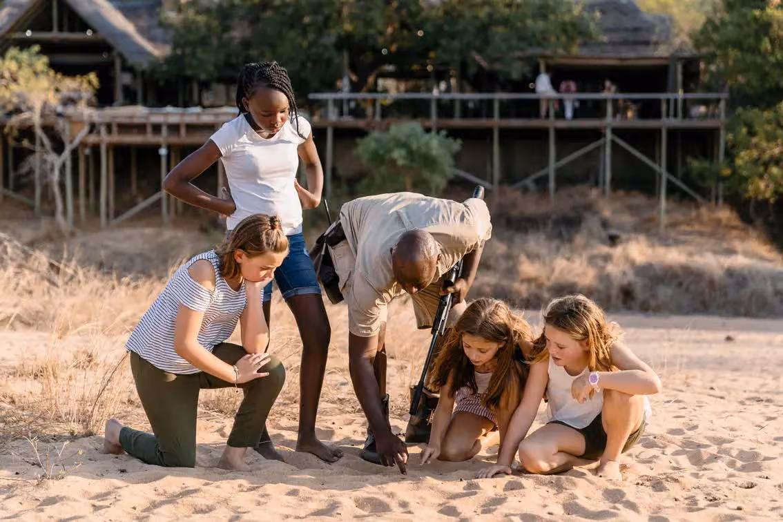 Man with rifle shows animal tracks in sand to four attentive children outdoors near a wooden structure.
