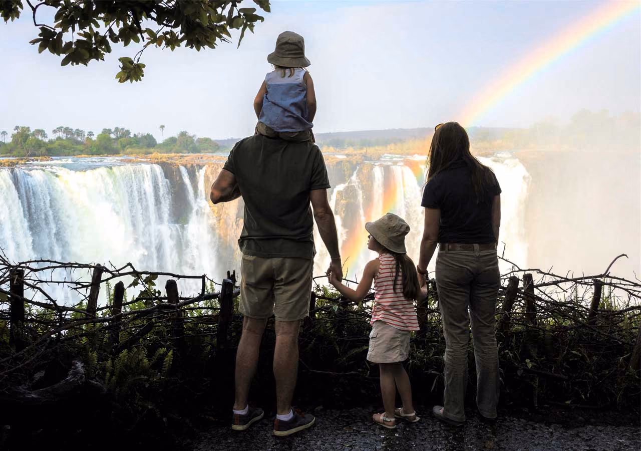 Family of four viewing large waterfall and rainbow over wooden fence in lush landscape.