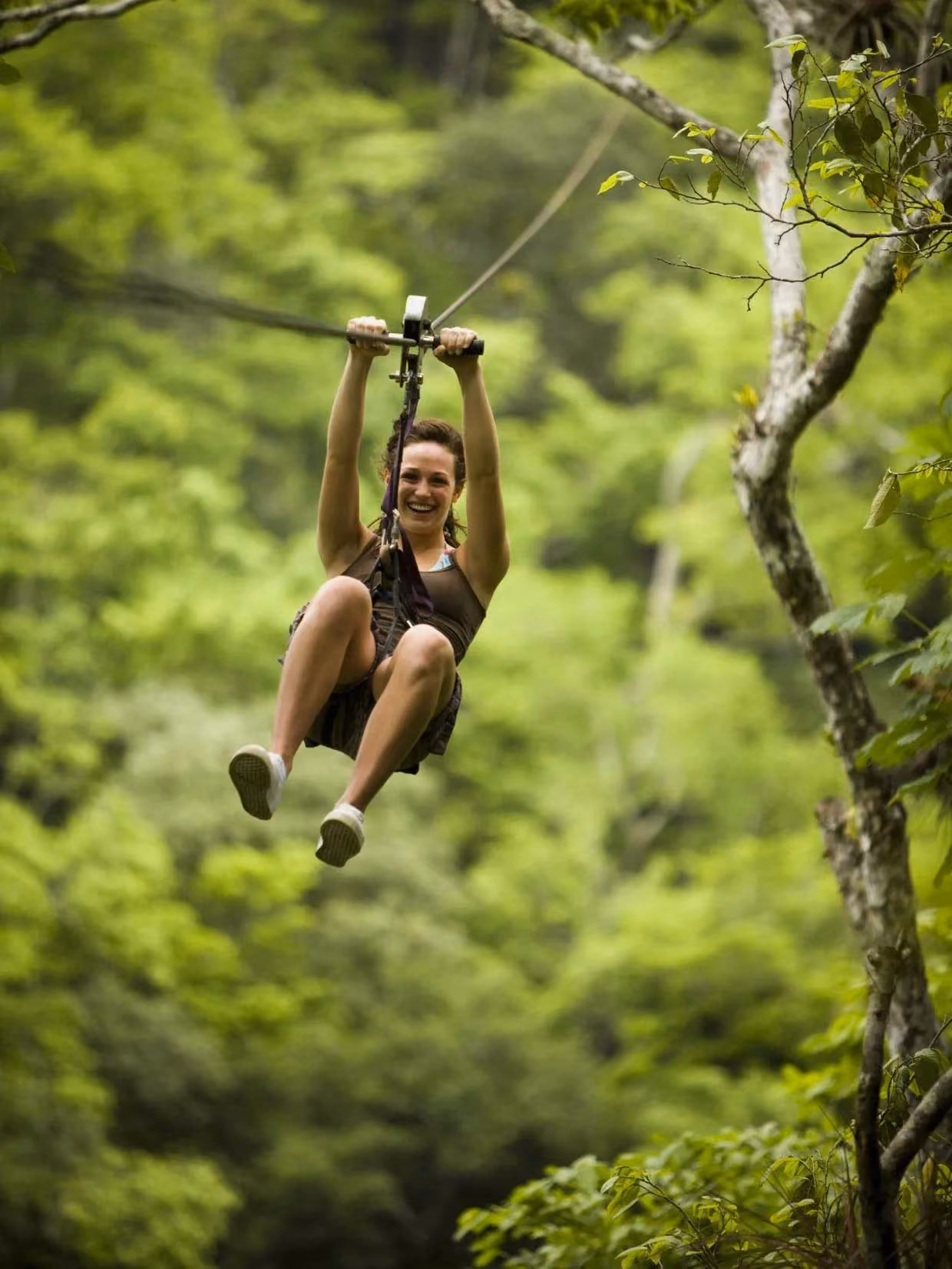Smiling woman zip-lining through dense, green forest wearing casual summer clothes and sneakers.