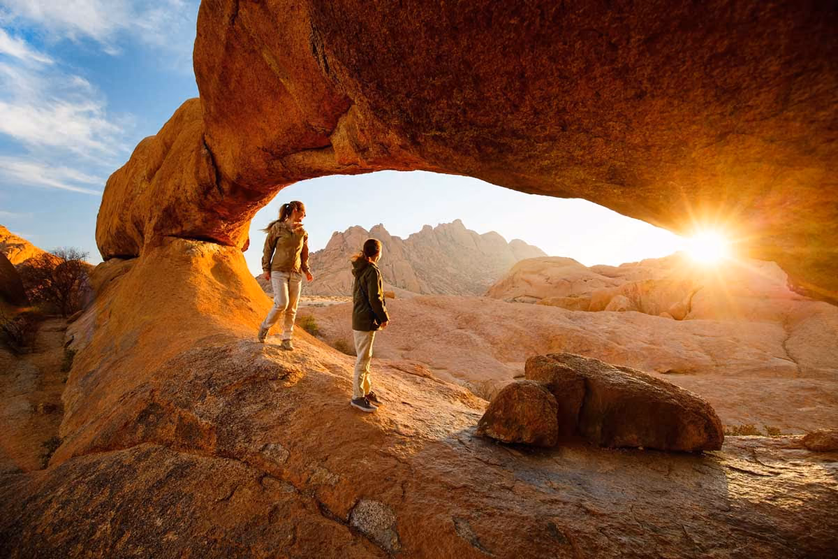 Two people standing on rocky terrain under a natural rock arch with the sun setting nearby.