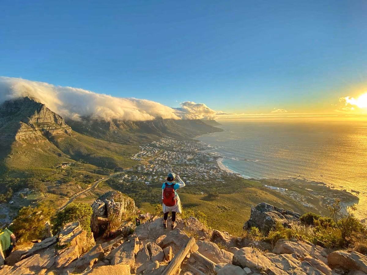 Person stands on rocky cliff overlooking coastal town, mountains wrapped in clouds, and ocean at sunset.
