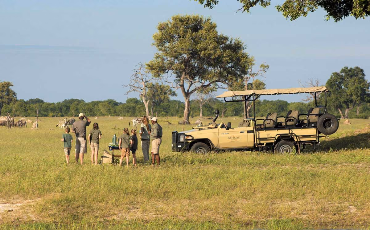 People on an African safari standing near an open vehicle watching zebras in a grassy savanna.